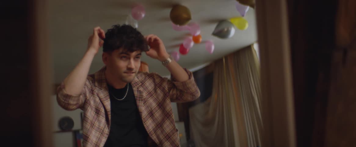 A man fixing his hair in a mirror while wearing a plaid suit with balloons on his ceiling