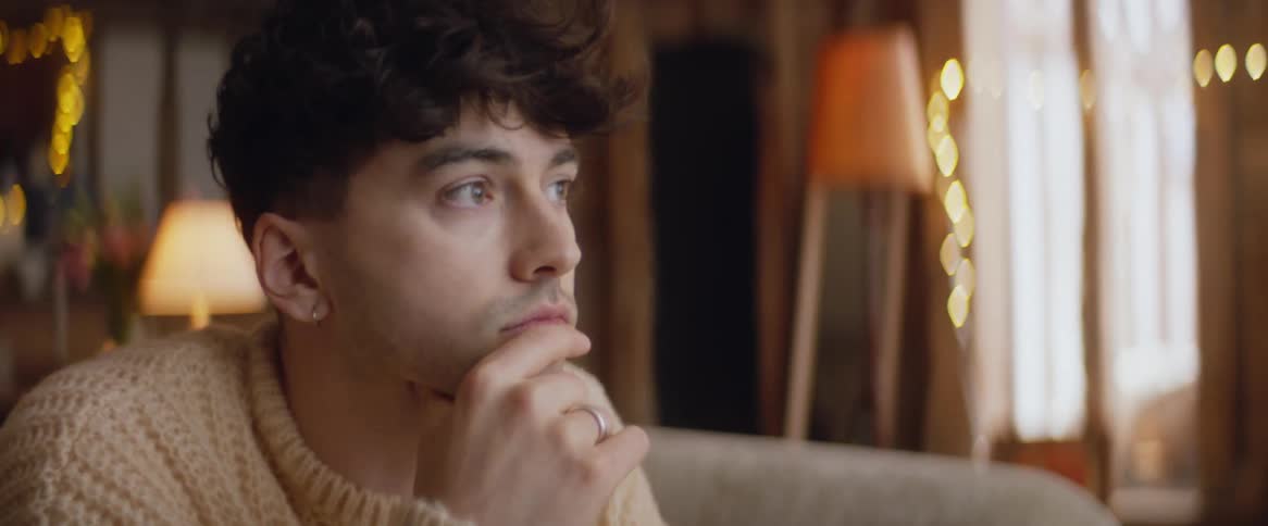 A man wearing a sweater sits on a couch with his hand on his face looking contemplatively