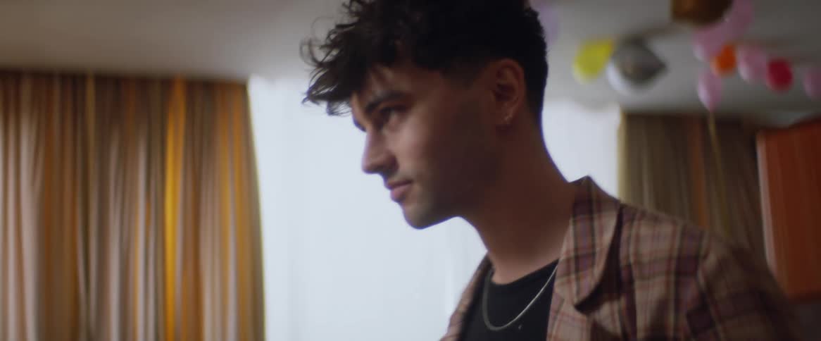 A male poses and looks at himself in a mirror with balloons on the ceiling in the background