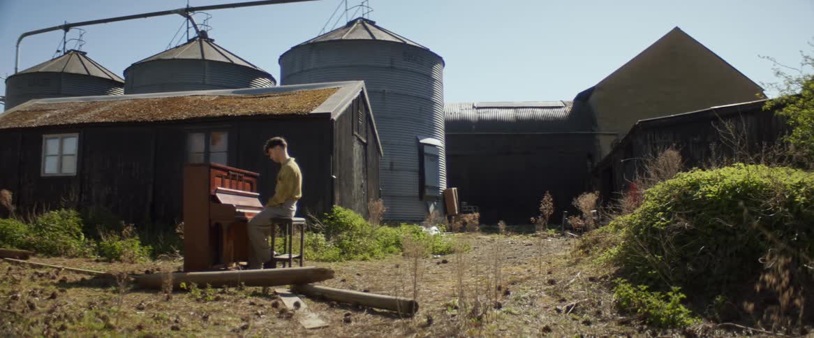 Man sits at piano outdoors in farm yard near grain silos