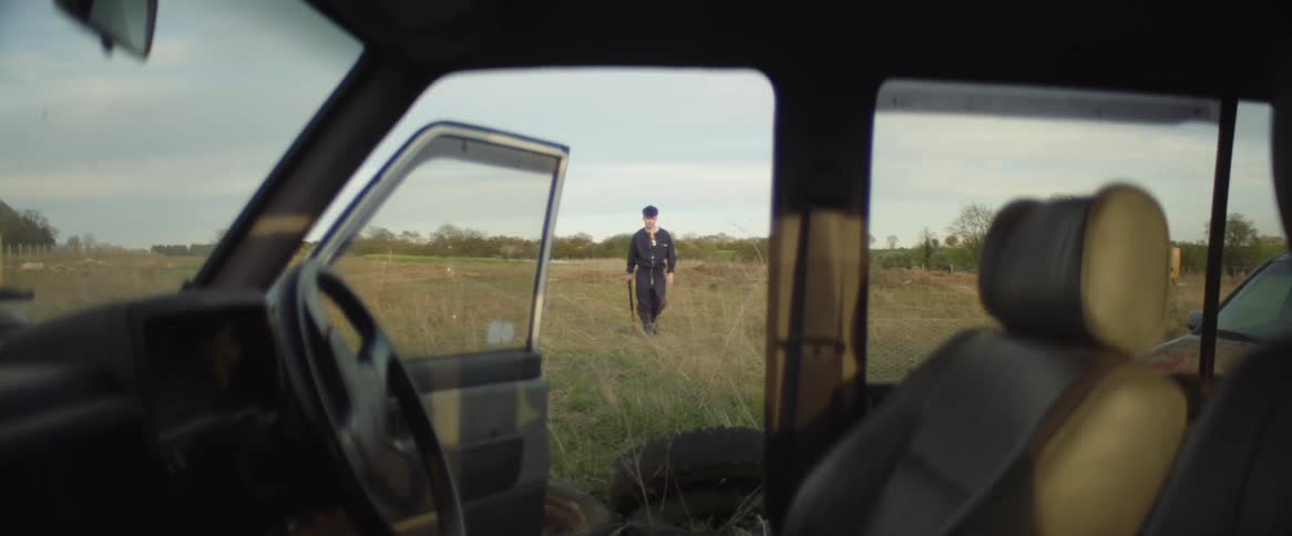 A man walks in the grass towards a car with its passenger side door open