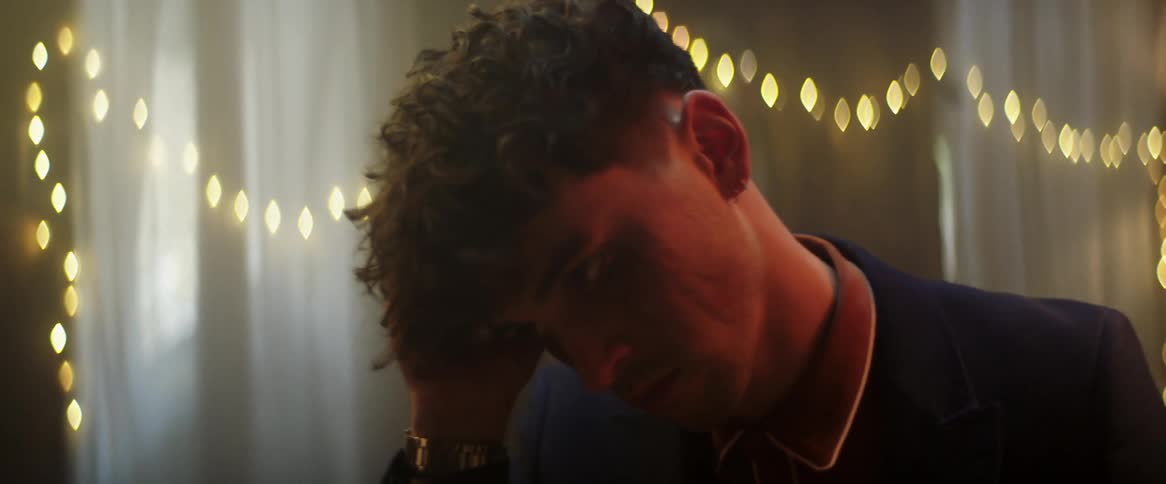 A teenage boy is standing inside a room rubbing the side of his head with his hand and he looks away