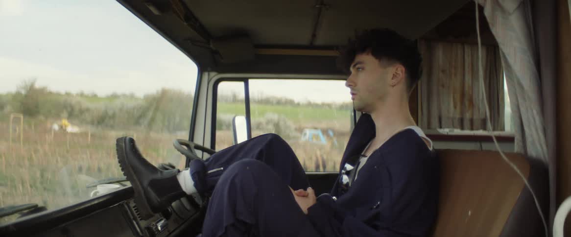 A teenage boy is sitting inside an old vehicle with his foot up near the steering wheel