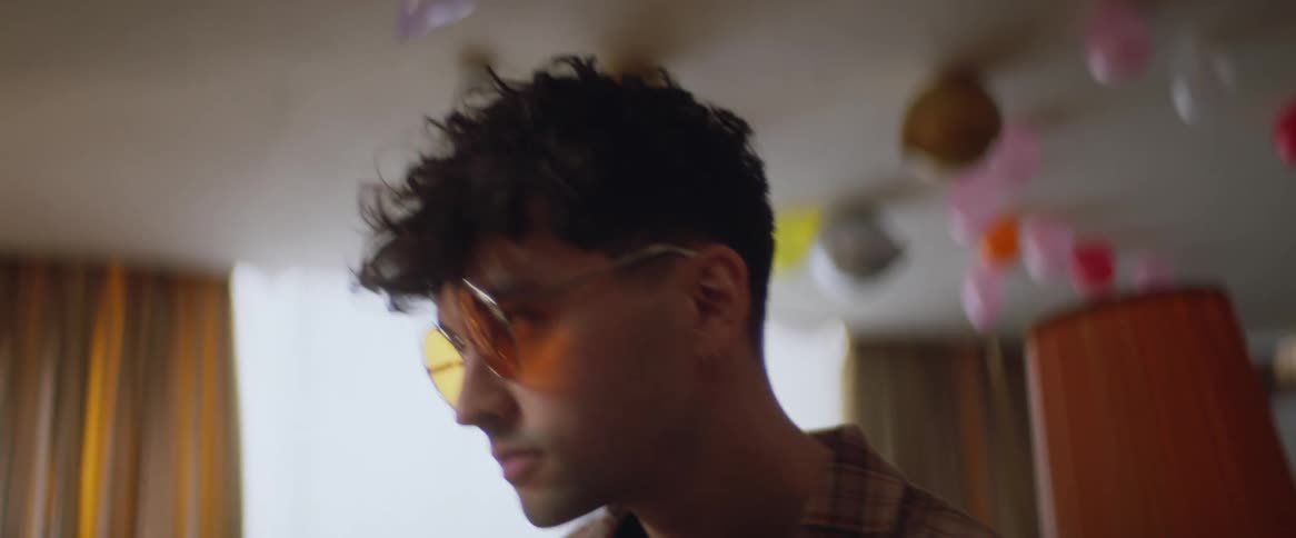 A man wearing glasses fixes his hair while looking in a mirror in a room with balloons on the ceiling