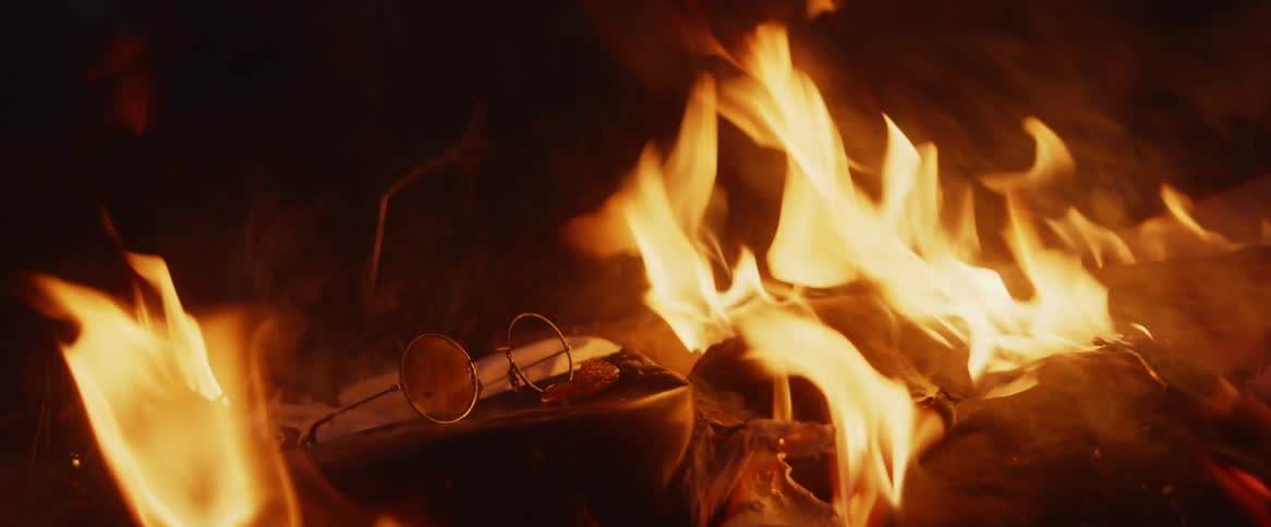 Round eyeglasses with one lens missing sit on pile of items burning at night