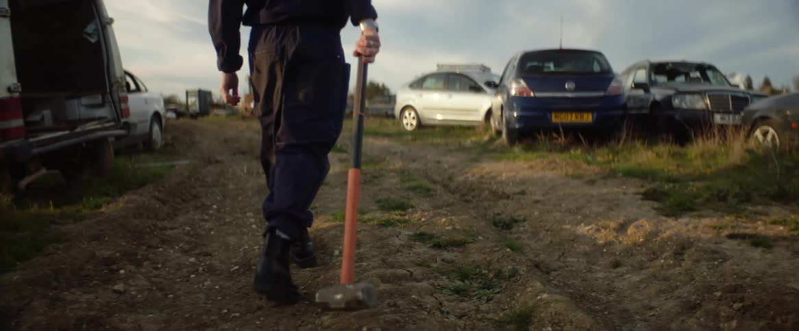 A man dragging a sledgehammer behind him is shown from behind walking into an area with a lot of parked cars