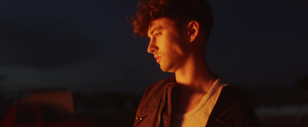 A teenage boy is standing outside watching a fire burn as the light shines on his face in the darkness
