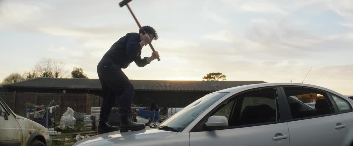 A young man is standing on the hood of a car swinging a sledgehammer as he breaks the windshield