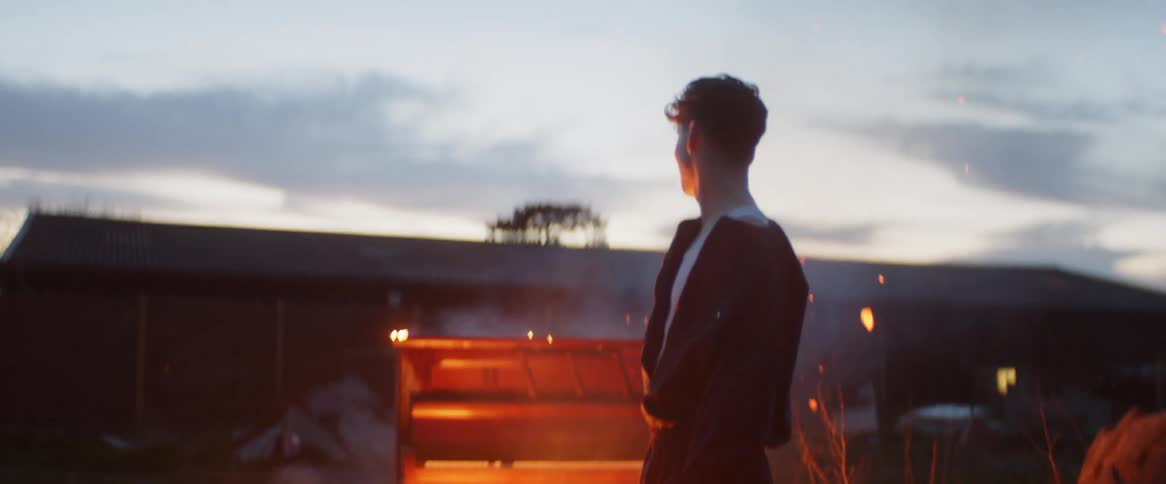 A man is standing in a barnyard in front of a piano that he has set on fire