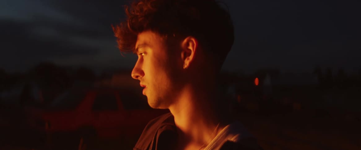 A teenage boy is standing outside watching a fire burn as the light shines on his face in the darkness