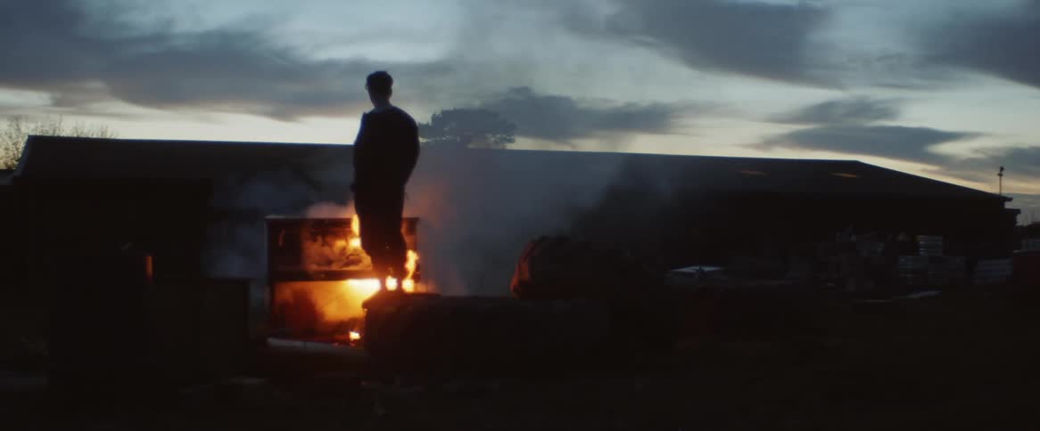 A silhouette of a man standing in front of a bonfire against a dusk sky