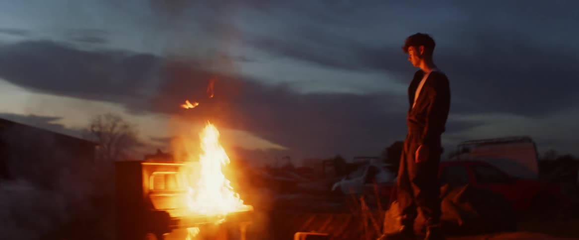 A teenage boy is standing outside on truck tires as he watches a piano burning in flames