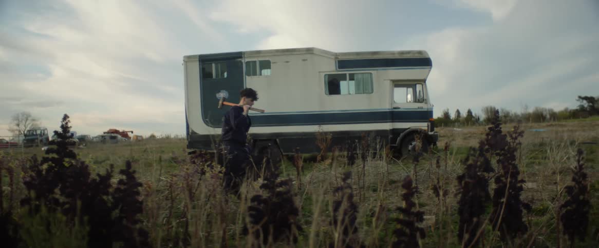 A man carries a sledgehammer through a field past a camper