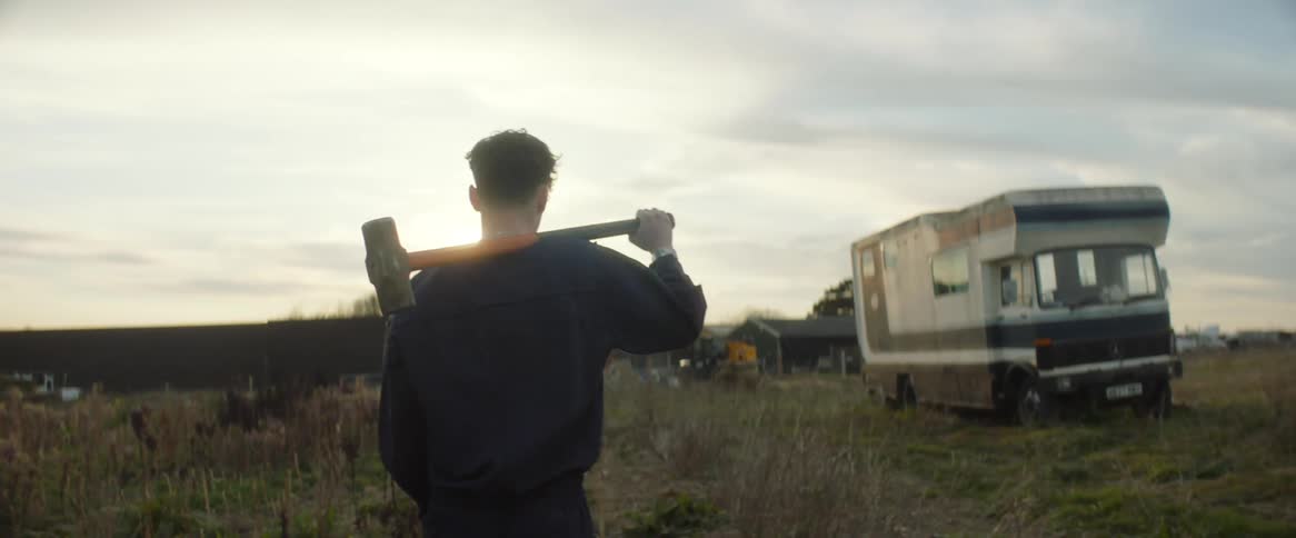 A man walking casually toward a camper and farm house with a sledge hammer over his shoulder