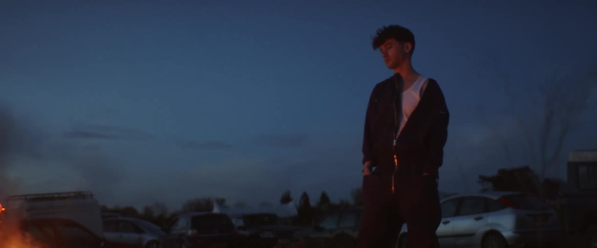 Man wearing a zip up jackets gazes into a campfire with a car parked behind him