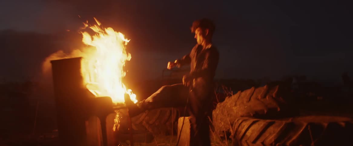 Man watching piano burn in field near tractor tires at night kicks the piano over