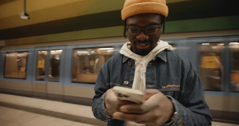A man laughs at his phone while standing with his back to a passing train