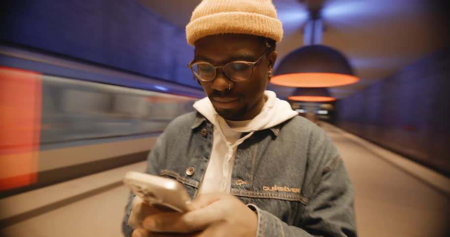 A man wearing a jacket and a knit cap is standing in the subway station texting on his phone as a train passes