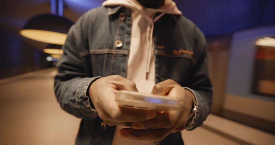 A man wearing a hoodie and a jacket is standing in the subway station texting on his phone with both hands as a train passes