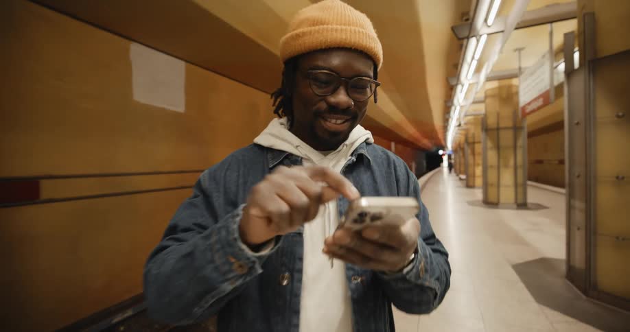 A young man standing in the subway station is smiling while clicking on his smartphone screen