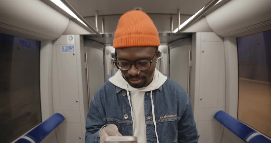 A man wearing a jacket and a knit cap is standing inside a moving subway train texting on his phone with his thumb