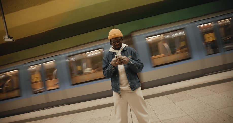 A man standing alone on a subway platform smiling and texting as a train passes behind him