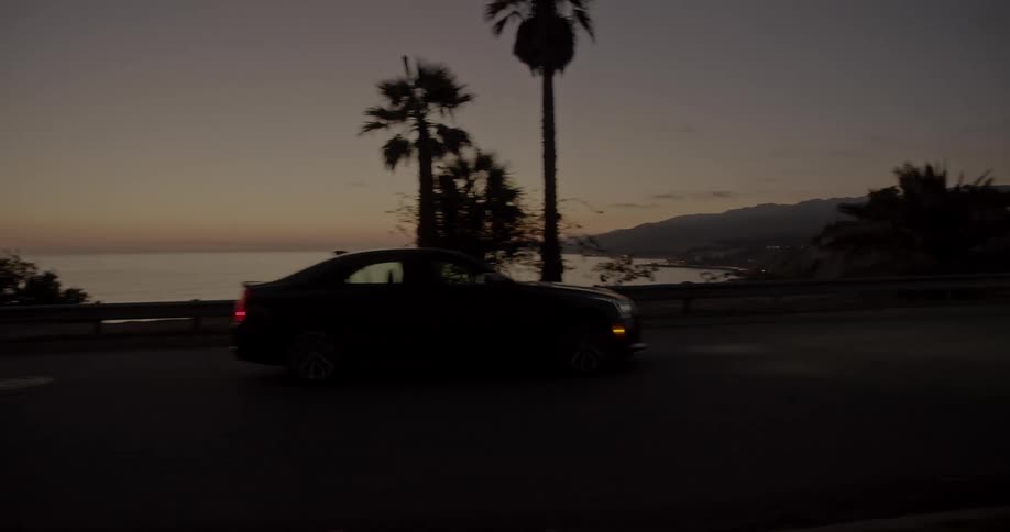 Car drives along highway near beach passing trees silhouetted against a tinted horizon