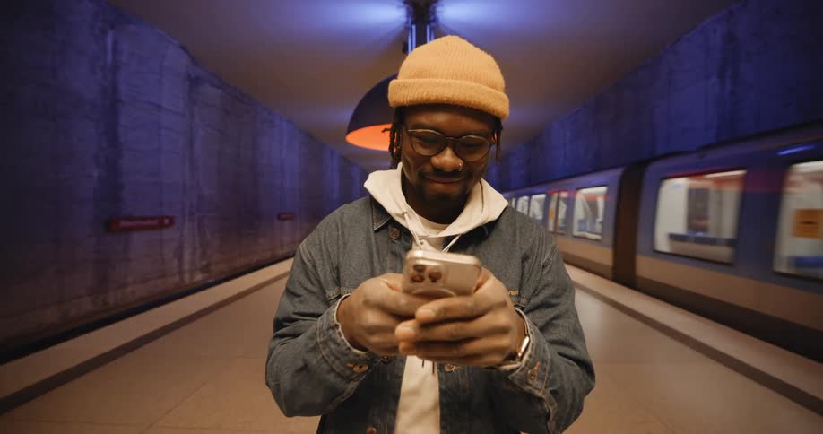 A man wearing a hoodie and a jacket is standing in the subway station texting on his phone with both hands as a train passes