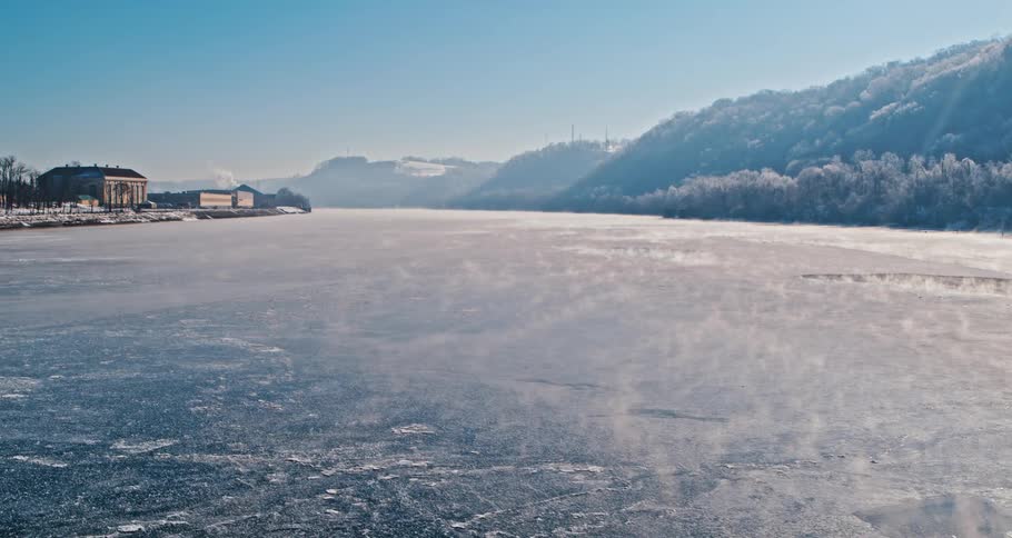 A frozen river lies between buildings and mountains under a clear sky on a winter day