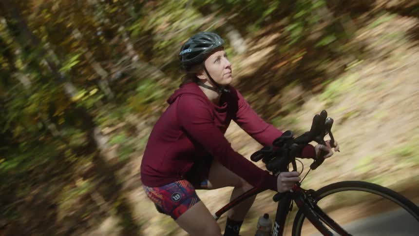 A woman wearing shorts and a jacket with a cycling helmet is riding her bicycle on a road near the woods
