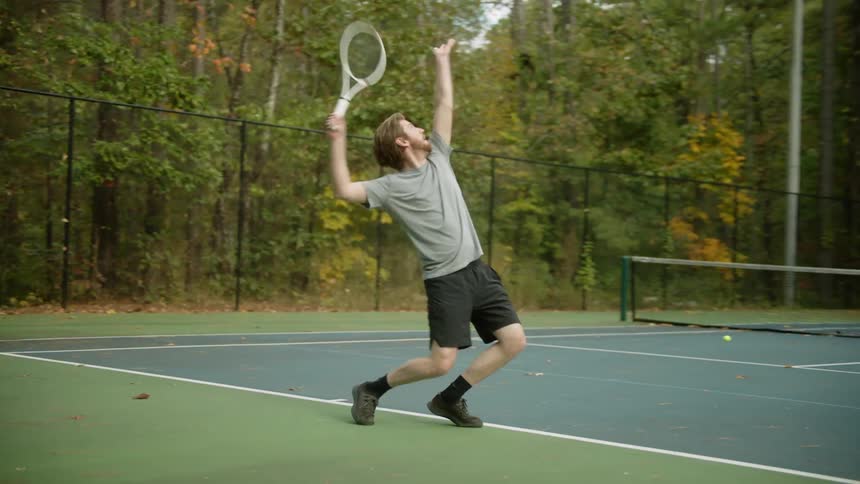 A man practices a serve on a tennis court before resting on a bench and drinking from a water bottle