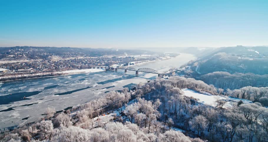 Snow covered trees are on the hills along the shoreline as a bridge crosses an ice overed river to the suburbs under a clear sky