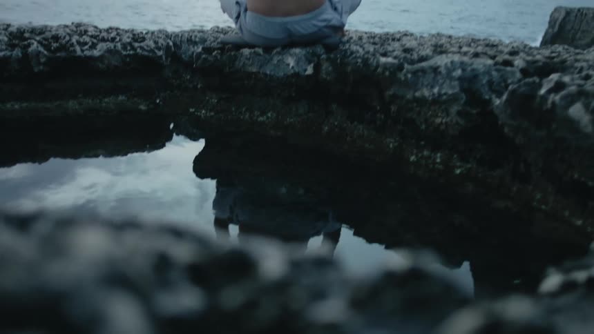 A man is sitting on rocks looking at the water as his body reflects on the water surface