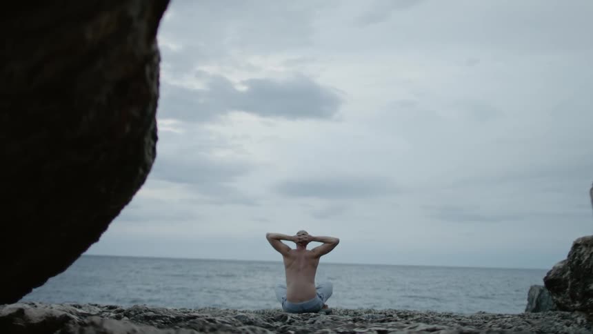 A man sitting cross legged on the beach stretching his hands behind his head