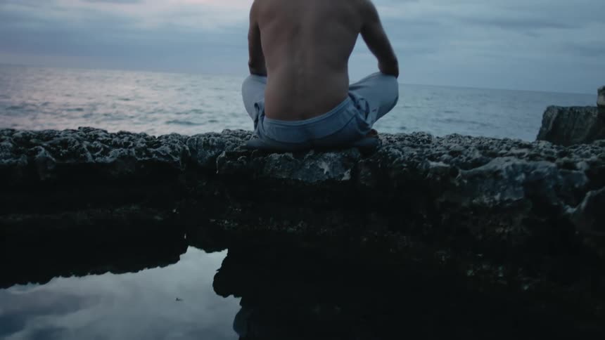 A man sits on a rocky shore and looks out at the ocean