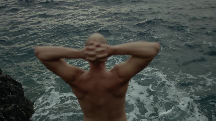 A man standing near the ocean stretching his hands behind his head