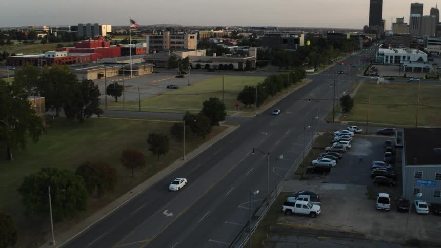 Light traffic moves on a city street with many buildings and parking lots and towers in the distance under a cloudy sky