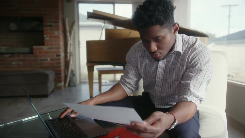 A man sitting typing on his laptop while looking at a paper