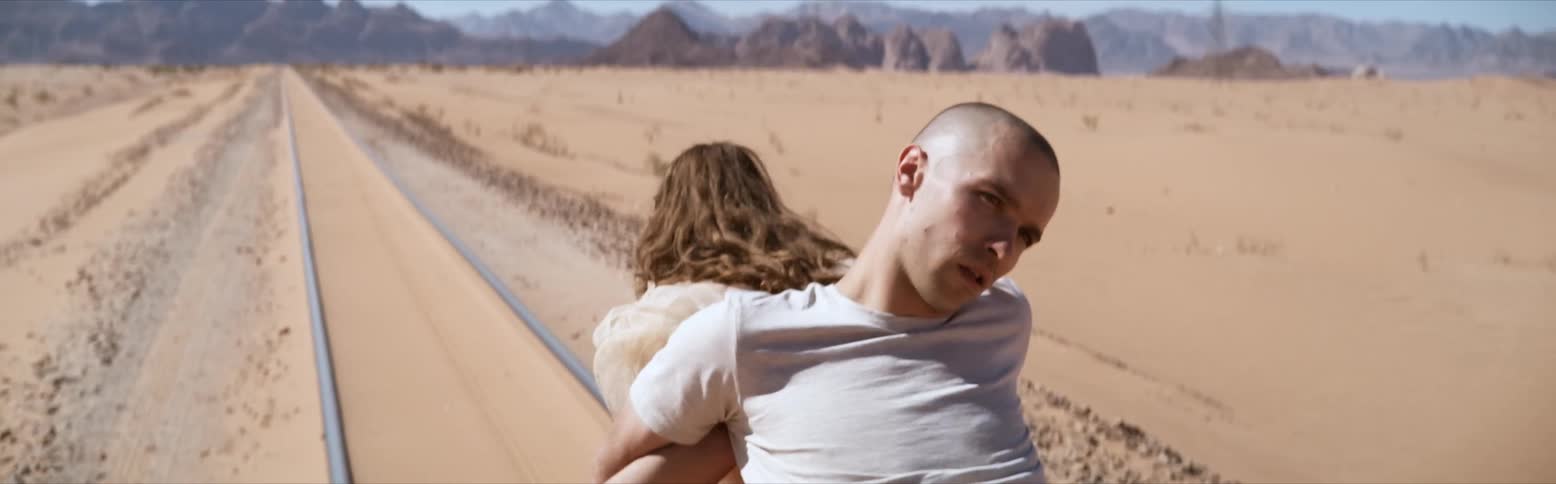 A woman carries a man on her back while walking through the desert