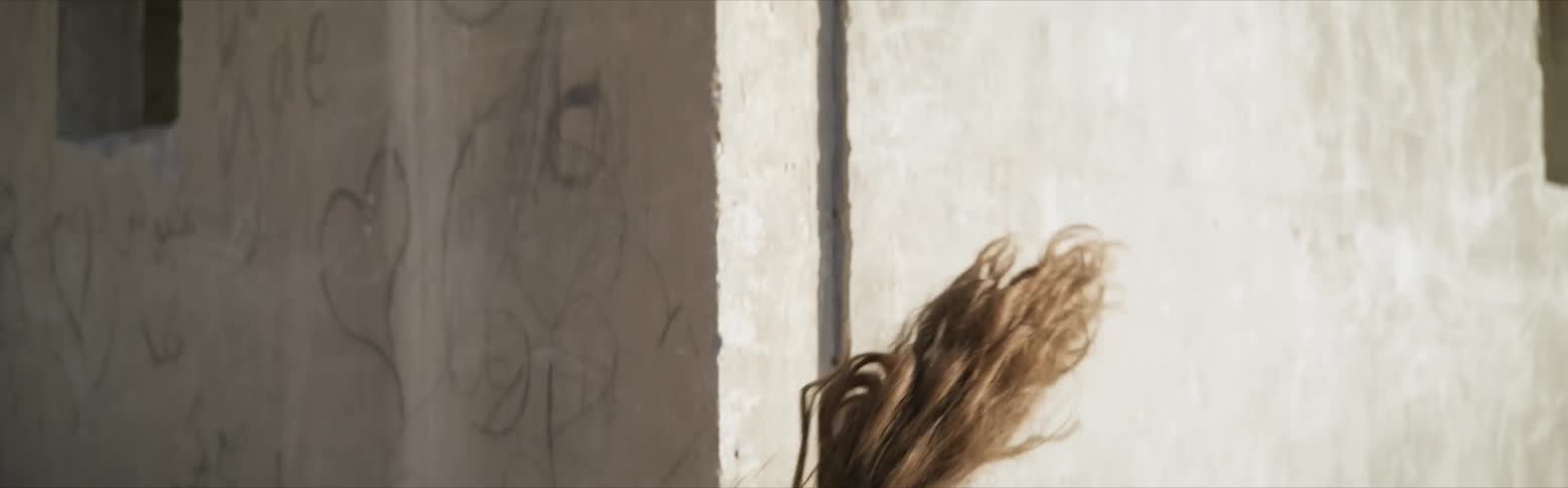 Young woman in seethrough flowy blouse dancing erratically up and down in and out of frame in front of concrete building