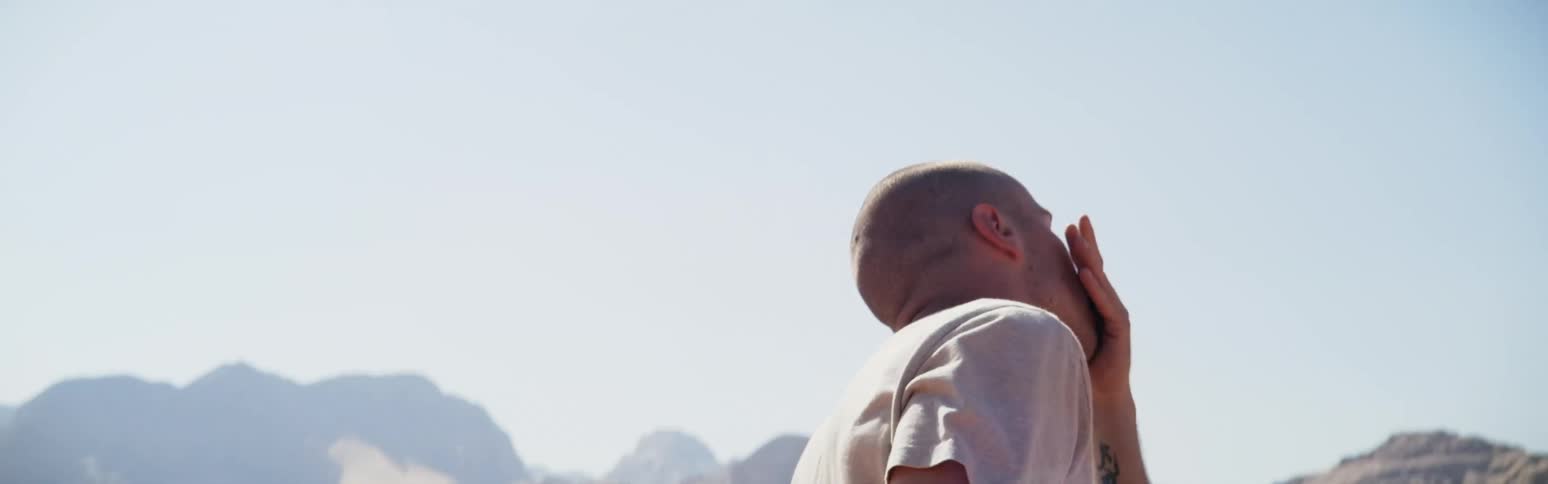 A man is standing in the desert dancing with his arms out as he turns and moves his hands around his face and head and there are mountains in the distance