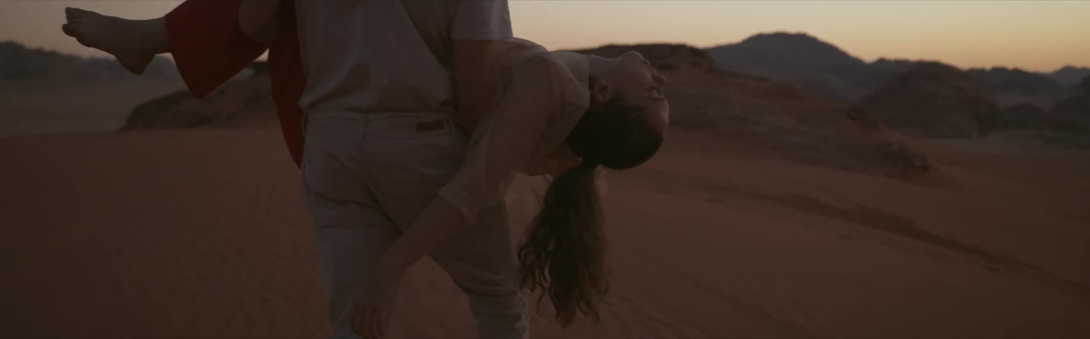 A man carrying a female in his arms across desert sand as she lays limp