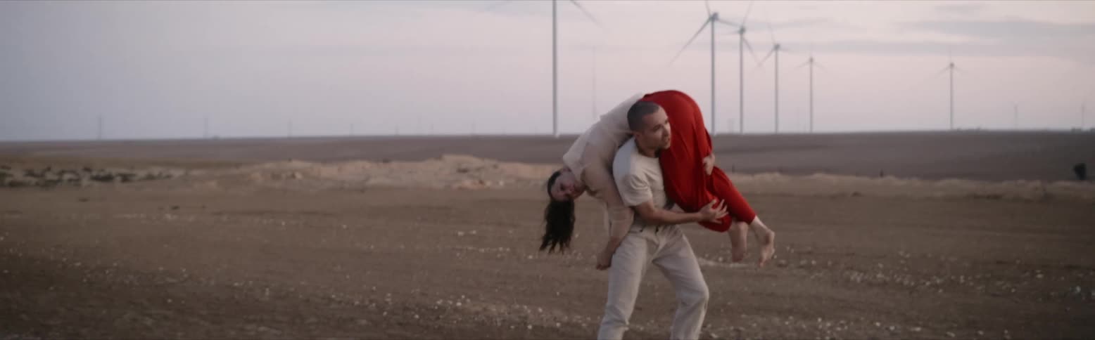 Man carries woman over his shoulder near road with wind farm in the distance