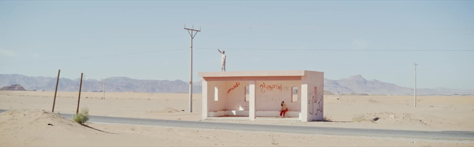 A building on the side of the road in the desert with a person sitting inside and another person on top