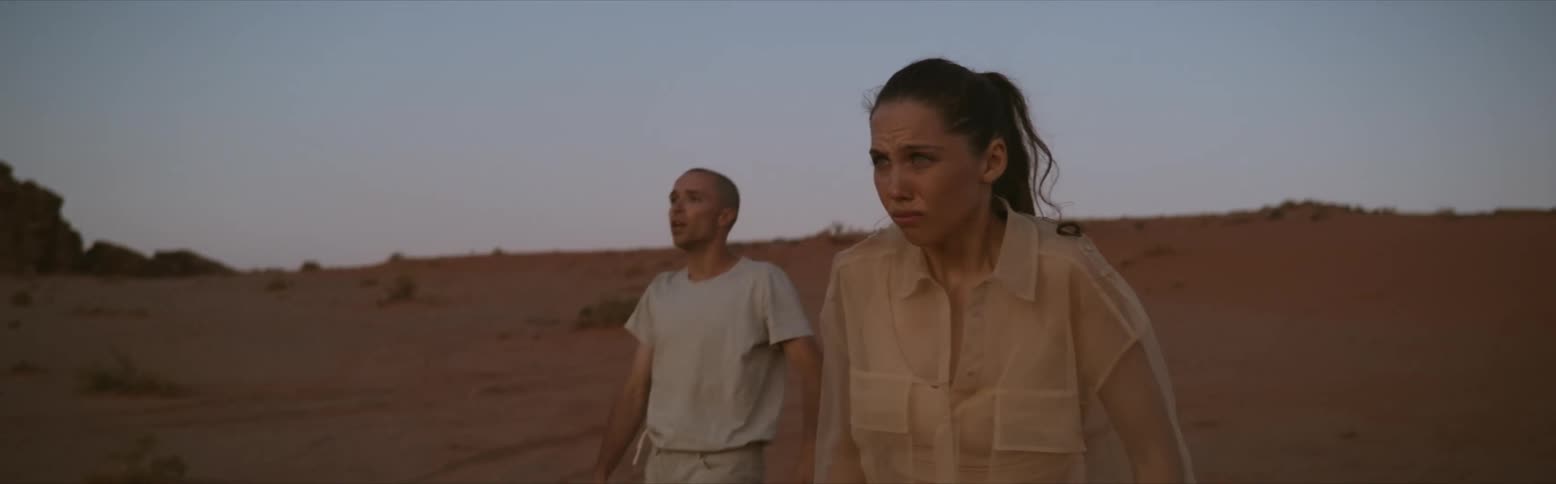 A terrified man and woman are struggling to run across the desert and they stop and look up