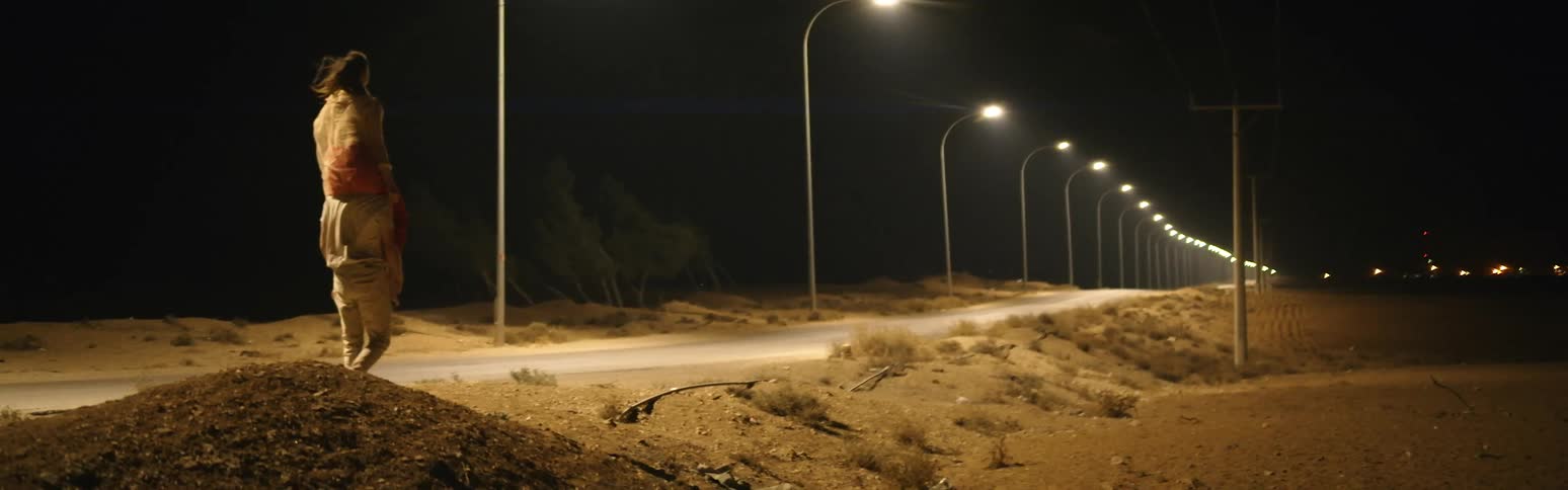 A woman is sitting on a mans shoulders as he walks carrying her on an empty desert road under the street lights at night