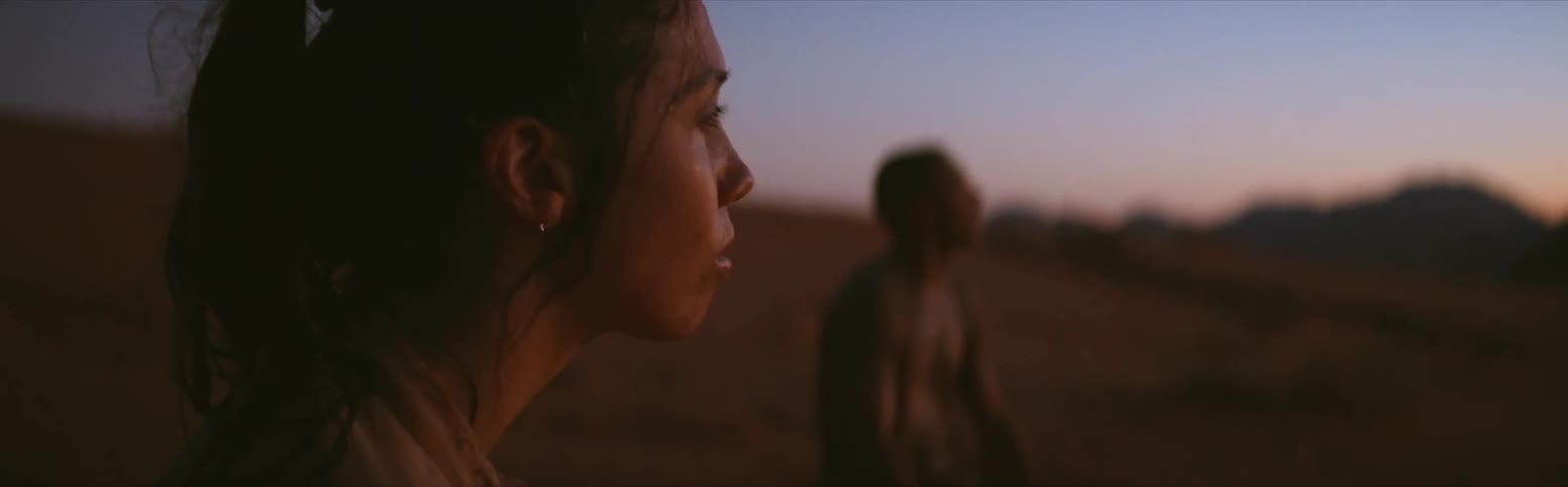 A woman is standing in the desert as she looks tired and there is a bare chested man near her