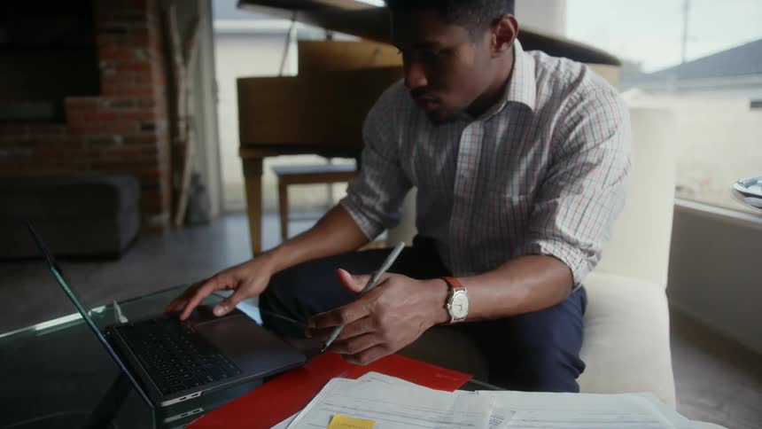 A man sits at home and looks at over due bills as he uses his laptop