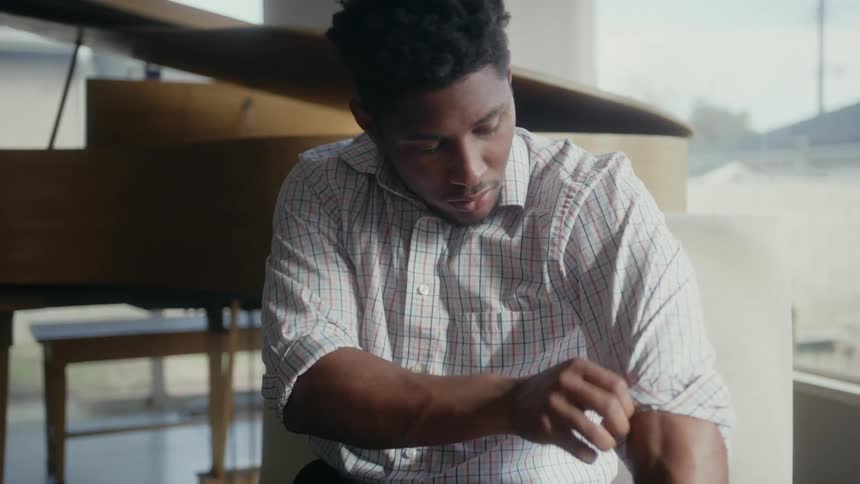 A man is sitting in a chair rolling up the sleeves of his shirt and there is a grand piano behind him