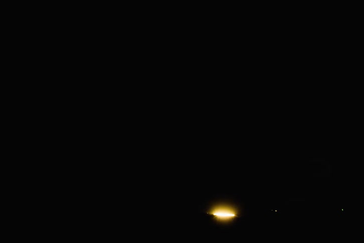 Lightning flashes brightly over mountains in a dark sky as car lights shine across the road